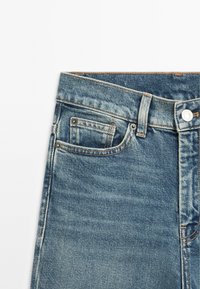 Jean en denim bleu avec une finition usée, comportant une petite poche avant, des accessoires en argent et des coutures contrastantes le long de la taille.