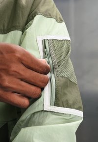 Hand holding the zipper of a green and olive jacket with mesh and white trim on the sleeve.
