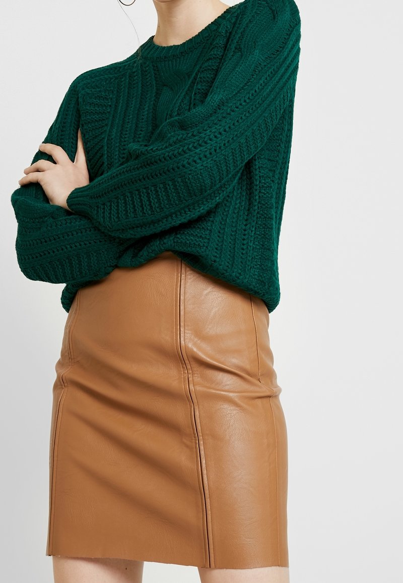 Femme portant un pull vert à maille torsadée à manches longues, rentré dans une mini-jupe en cuir marron clair, les bras croisés.