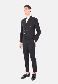 Completo doppiopetto nero con bottoni dorati, caratterizzato da un taglio su misura, tasche laterali e una camicia bianca sottostante. Accompagnato da mocassini neri.