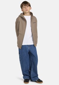 Felpa beige con zip e tasche frontali, indossata sopra una t-shirt bianca. Accoppiata con jeans blu larghi e sneakers nere. Logo nero semplice sulla felpa.