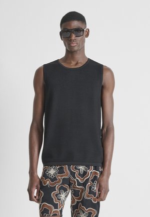 Man in mouwloze zwarte top, grote zwarte zonnebril en zwarte broek met bruin-witte bloemenpatronen, staand tegen een effen achtergrond.