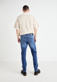 Beige kortärmad skjorta, blå denimjeans med en urtvättad finish, och svarta skor med kraftig sula, sett bakifrån.