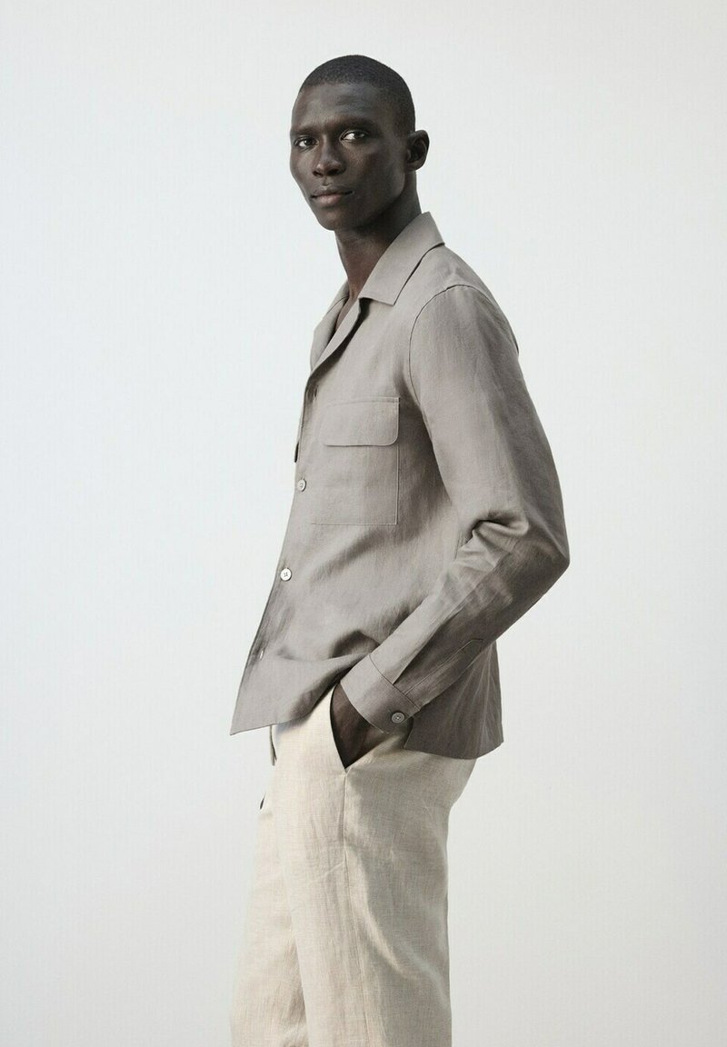 Modèle masculin portant une chemise boutonnée marron clair et un pantalon beige, debout de profil, les mains dans les poches, sur un fond gris uni.