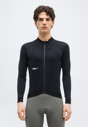 Νέος άνδρας που φοράει εφαρμοστό μαύρο μακρυμάνικο ποδηλατικό φανελάκι και γκρι επενδυμένα ποδηλατικά σορτς, στέκεται μπροστά σε απλό φόντο.