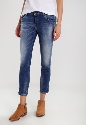 Person trägt ausgewaschene blaue Skinny-Jeans, weißes Hemd und braune Wildleder-Stiefeletten und steht auf hellem Hintergrund.