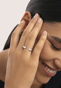 Silberne Ringe mit einem glatten, ovalen Design werden an den Fingern einer Hand getragen, die das Gesicht berührt. Die Haut hat einen warmen Farbton; die Nägel sind hellrosa.