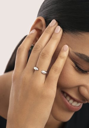 Anillos de plata con un diseño ovalado y suave se llevan en los dedos de una mano que toca la cara. La piel tiene un tono cálido; las uñas son de un rosa claro.