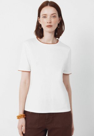Frau mit schulterlangem braunem Haar, die ein weißes Kurzarmshirt mit braunem Rand und braune Hosen trägt, accessoriert mit einem Bernsteinarmband und Creolen.