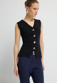 Czarny bezrękawnik z dekoltem w kształcie litery V, z pięcioma złotymi guzikami z przodu i lekko rozszerzanym dołem. Połączony z granatowymi spodniami.