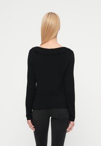 Γυναίκα με ίσια ξανθά μαλλιά, που φοράει μαύρο μακρυμάνικο τοπ με βολάν στον γιακά, εμφανιζόμενη από πίσω σε λευκό απλό φόντο.