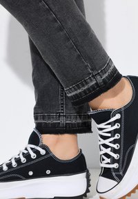 Zapatillas de lona negras con cordones blancos, con una media suela blanca y una suela exterior de goma negra texturizada, combinadas con jeans de mezclilla oscura.