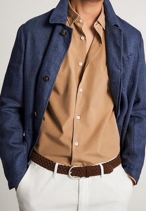 Homme portant un blazer bleu sur une chemise beige à boutons, un pantalon blanc et une ceinture tressée marron, les mains dans les poches.
