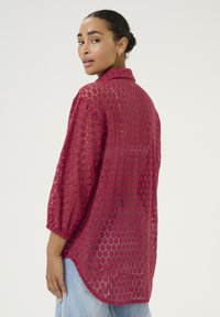 Lace blouse in deep burgundy with a hexagonal pattern, three-quarter sleeves, and a relaxed fit, featuring a curved hem and collar.