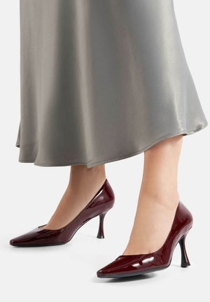 Tacones altos de charol burdeos con punta puntiaguda, tacón delgado y detalle recortado en la parte superior, combinados con un vestido de satén gris.