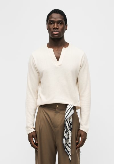 Άνδρας που φοράει κρεμ μακρυμάνικο μπλουζάκι με V λαιμόκοψη και καφέ παντελόνι, με ένα ασπρόμαυρο φουλάρι με σχέδιο βαλμένο στη μέση του παντελονιού.