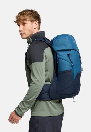 Zaino da trekking blu e blu scuro con spallacci regolabili, tessuto strutturato e chiusura superiore. Indossato da una persona con abbigliamento verde e nero.