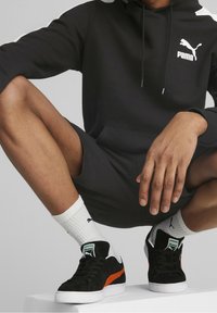 Felpa nera Puma con logo bianco, abbinata a scarpe da ginnastica in suede nero con striscia arancione e suola bianca, e calzini bianchi alla caviglia.