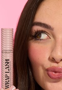 Vaaleanpunainen ripsiväriputki, jossa on tekstissä "WRAP LASH" ja aaltoileva muotoilu, naisen kasvojen vieressä, jolla on tarkasti määritellyt ripset ja kiiltävät huulet, vaaleanpunaisen taustan edessä.