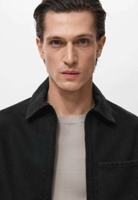 Črna jakna na zadrgo s ovratnikom, kombinirana s svetlo sivo majico. Gladko teksturo tkanine, minimalističen dizajn. Nevtralna ozadje.