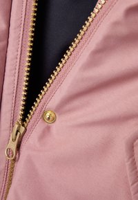 Tessuto rosa con una texture liscia, caratterizzato da una cerniera dorata e un bottone a scatto. Primo piano dell'area con cerniera su uno sfondo scuro.