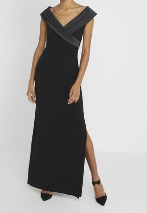 Femme debout portant une robe de soirée noire sans manches avec un décolleté en V profond et une fente latérale, assortie à des chaussures noires pointues.