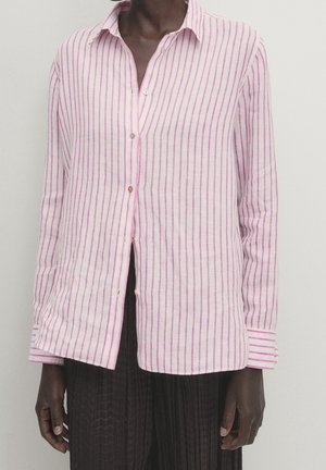 Personne portant une chemise blanche à manches longues avec des rayures verticales roses et un pantalon plissé noir sur un fond neutre.