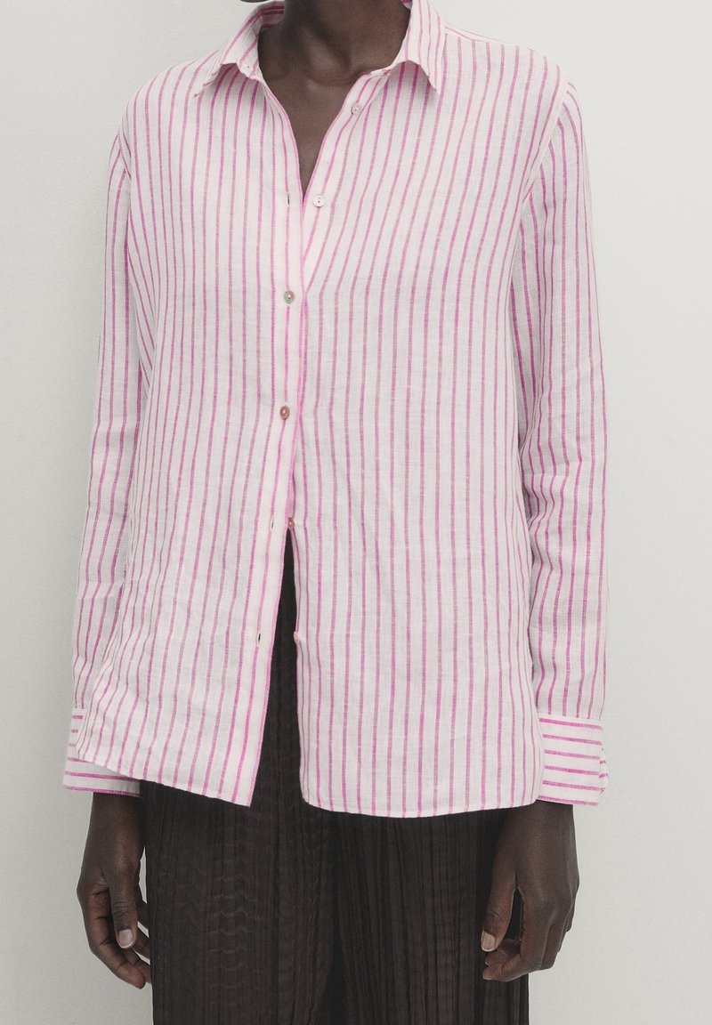 Personne portant une chemise blanche à manches longues avec des rayures verticales roses et un pantalon plissé noir sur un fond neutre.