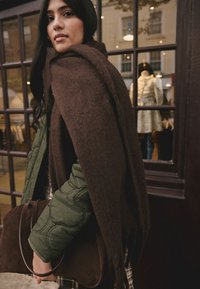 Brown oversized shawl draped over a green quilted jacket. The individual is holding a brown suede bag. Background features store window.
