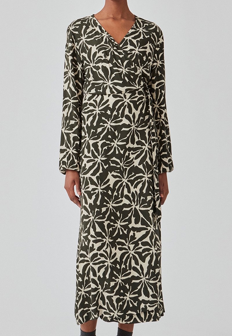 Femme portant une robe portefeuille à manches longues, longueur cheville, avec un motif abstrait de feuilles noir et blanc sur fond clair.