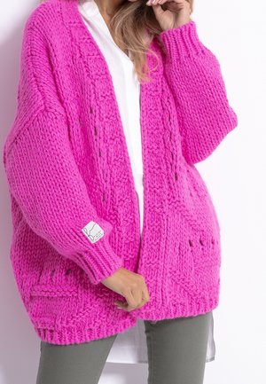 Kobieta w jaskrawo różowym, grubym kardiganie z warkoczowymi wzorami, nałożonym na białą koszulę i szare spodnie, trzymająca dół kardiganu.