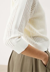 Biały dzianinowy sweter z otwartym wzorem splotu, ściągaczami na mankietach i luźnym krojem. Zdjęcie przedstawia widok z boku z widoczną miękką fakturą.