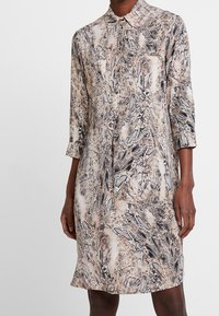 Kobieta w sukience do kolan o beżowym i czarnym wzorze w węża oraz rękawach trzyczworćowych na jednolitym tle.