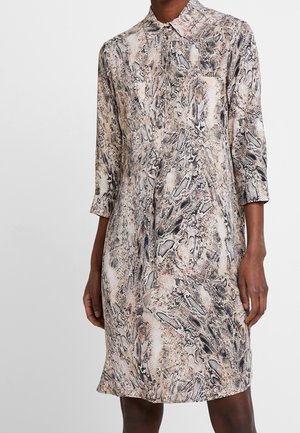 Vrouw in een knielang jurkje met een beige en zwart slangenprint en driekwartmouwen voor een effen achtergrond.