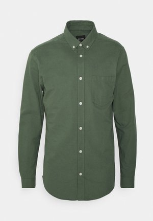 Chemise en coton verte à manches longues avec col boutonné, poche frontale et boutons blancs. Texture lisse et design d'ourlet incurvé.