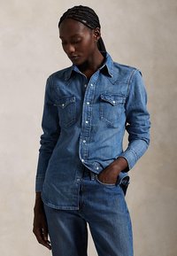 Frau mit geflochtenem Haar, die ein langärmeliges Jeanshemd und Jeans trägt, mit einer Hand in der Tasche und nach unten blickt.