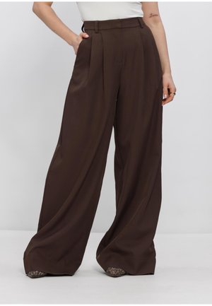 Frau trägt hochgeschnittene, weit geschnittene dunkelbraune Hose mit Seitentaschen, kombiniert mit einem weißen Oberteil, steht vor einem einfarbigen Hintergrund.