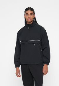 Giacca nera a pullover con colletto alto, zip frontale a metà, polsini elastici e una striscia bianca a contrasto lungo il petto; realizzata in tessuto leggero.