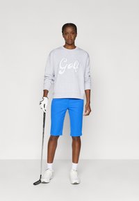 Grå hettegenser med "Golf" i hvit skrift, kombinert med lyseblå shorts. Hvite joggesko og en golfklubbe i hånden.