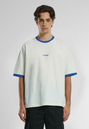 T-shirt bianco oversize con colletto e polsini a costine blu. Presenta la scritta "Proibito" ricamata in blu vicino al petto. Tessuto liscio.
