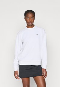 Biely sveter s okrúhlym výstrihom, rebrovanými lemami a malým zlatým logom na hrudi, v kombinácii s čiernou športovou sukňou.