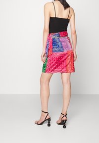 Mini-jurk met kleurrijke bandana-print, bestaande uit roze, blauw, groen en rode secties, met een gestrikte taille; model draagt zwarte sandaaltjes met bandjes.
