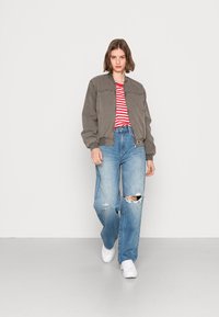 Casaco bomber cinza por cima de uma t-shirt riscada em vermelho e branco, combinado com jeans claros com efeito desgastado e ténis brancos.