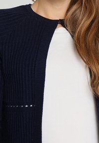 Mörkblå ribbad cardigan med figurnära design, rund halsringning och liten knappstängning, bärs över en vit topp.