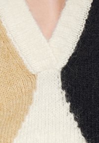 Sweter dziewiarski z dekoltem w kształcie litery V, wykonany z beżowej, kremowej i czarnej włóczki, z teksturowanym wzorem i widocznym szyciem.