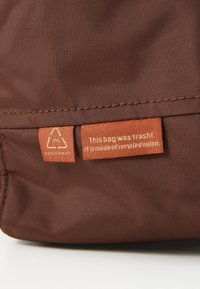 Brun resirkulert nylonveske med teksturert overflate og en oransje label som sier: "Denne vesken var søppel! Den er laget av resirkulert nylon."