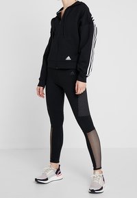 Felpa nera con zip e strisce bianche, abbinata a leggings neri con pannelli in rete. Sneakers bianche e rosa completano l'outfit.