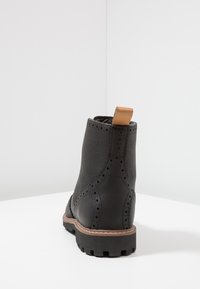 Stivale alla caviglia in pelle nera con dettagli brogue, targhetta di estrazione color tan e finitura testurizzata. Suola in gomma con un modello di battistrada visibile.