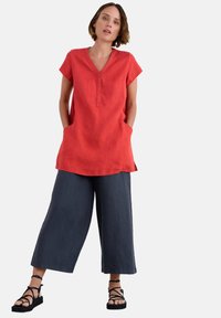Rode linnen tuniek met een V-hals en korte mouwen, voorzien van een knopenlijst. Gepaard met losse, donkergrijze cropped broek en gespsandalen in het zwart.