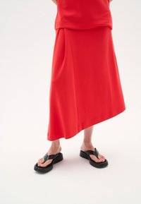 Rotes Strickoberteil und asymmetrischer Midirock; schwarze Plateau-Zehentrenner. Glatte Textur, schlichtes Design und kontrastreiche Farbelemente.